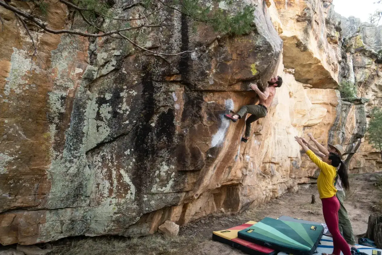 Ein Mann klettert an einer Felswand, w&auml;hrend zwei Personen unten sichern. Das ist was ist bouldern: Klettern ohne Seil an niedrigen Felsen mit Matten zur Absicherung.