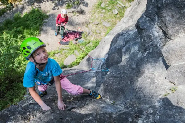 Ein Mädchen mit Helm klettert an einem Felsen, während eine andere Person sichert. Ein tolles Erlebnis beim Klettern mit Kindern.