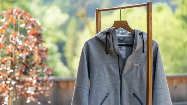 Graue Lodenjacke mit Kapuze, Reißverschluss und Taschen, aufgehängt an einem Holzständer vor blühendem Baum und grüner Natur.